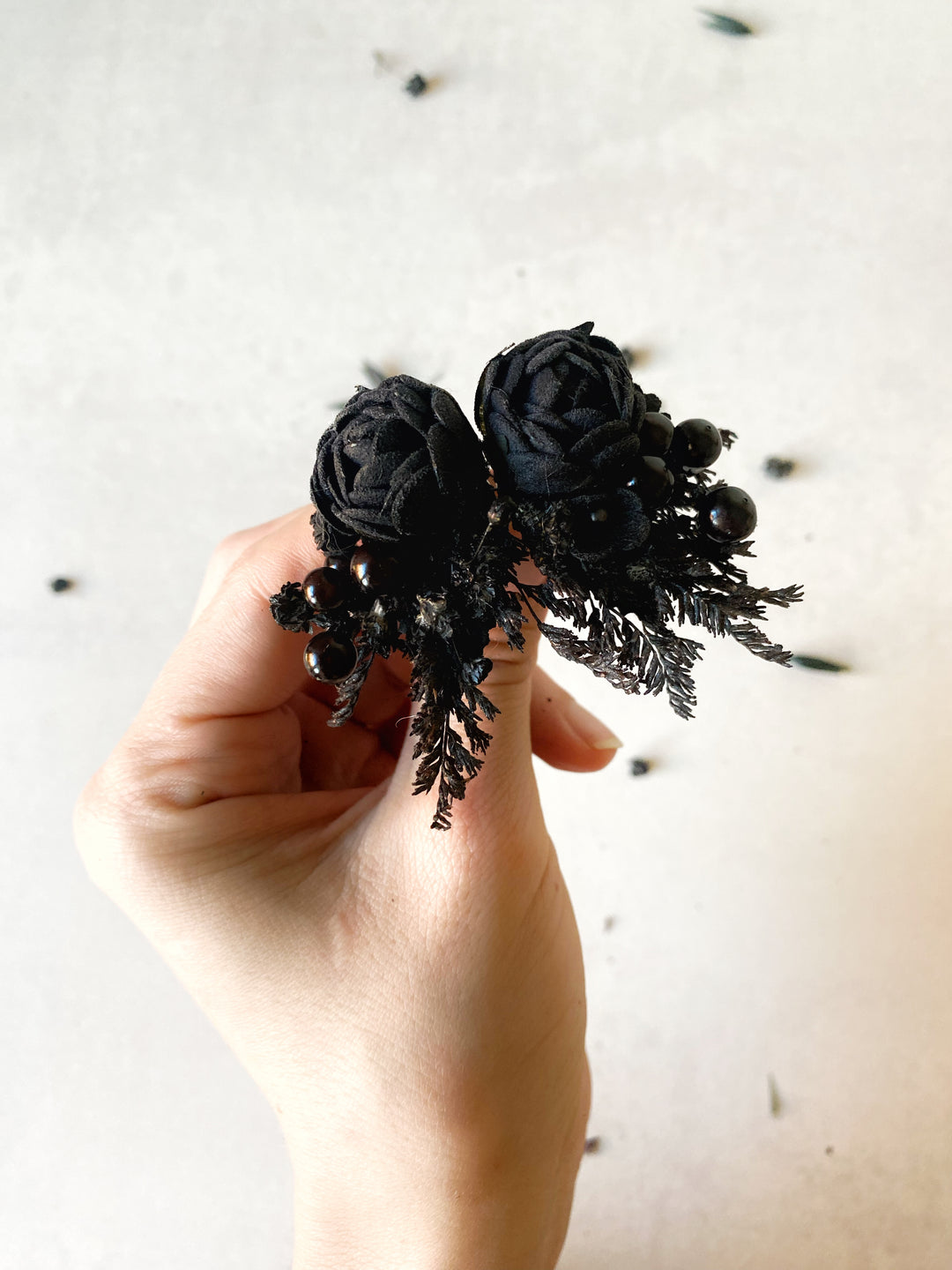 A hand holding a black floral earrings with two dark roses, black beads, and textured black foliage against a light neutral background.