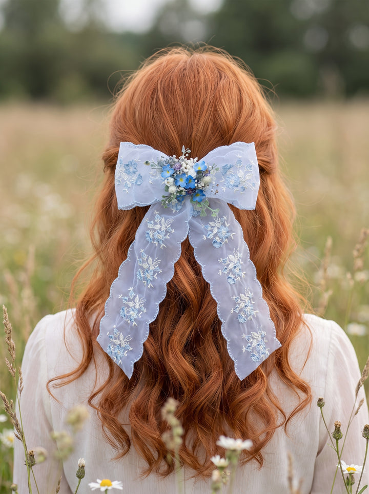 A person seen from behind wearing a large light blue lace hair bow with floral embellishments in a grassy outdoor field.
