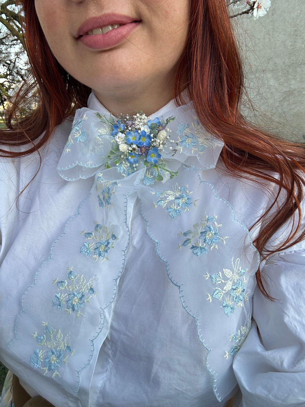 A close-up of a person wearing a white blouse with delicate blue and white floral embroidery and sheer ruffled collar details, shown outdoors with red hair visible.