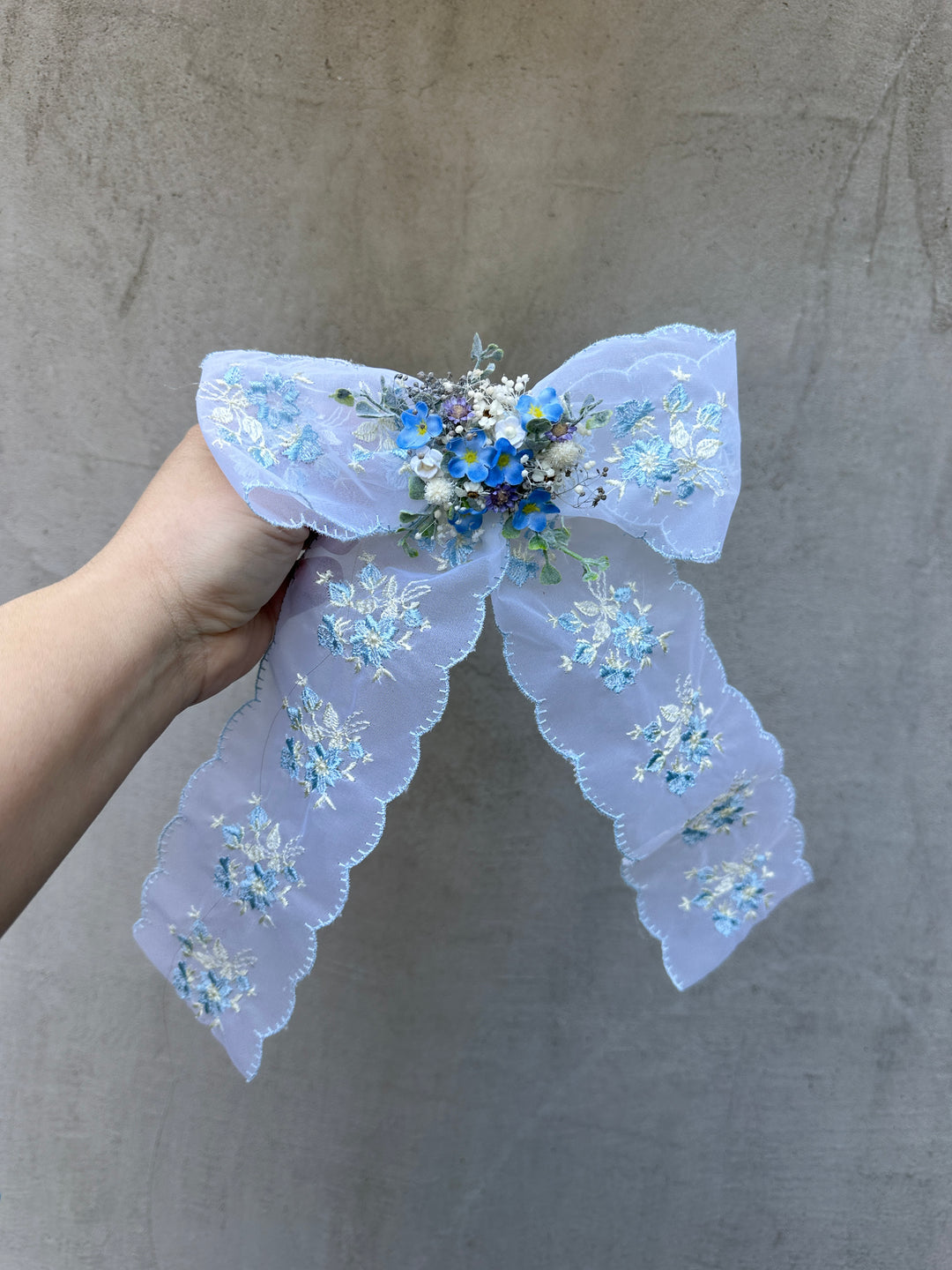 A hand holding a light blue lace hair bow decorated with embroidered flower patterns and a small blue floral center, set against a textured gray background.
