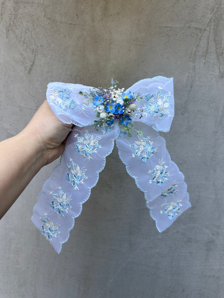 A hand holding a light blue lace hair bow decorated with embroidered flower patterns and a small blue floral center, set against a textured gray background.