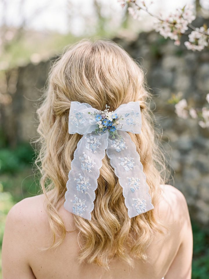 A blonde woman shown from behind wearing a sheer light blue lace hair bow accessory with small blue and white floral details, set against a soft-focus outdoor background with stone wall and blossoms.