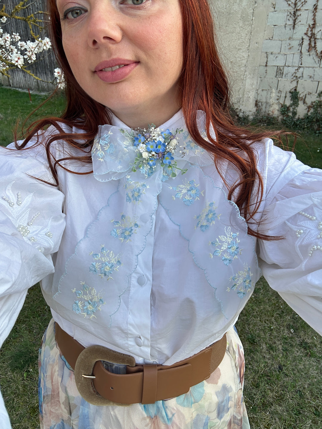 A woman wearing a white long-sleeve blouse with delicate blue and white floral embroidery, styled with a wide brown belt and a light floral skirt, photographed outdoors on grass.