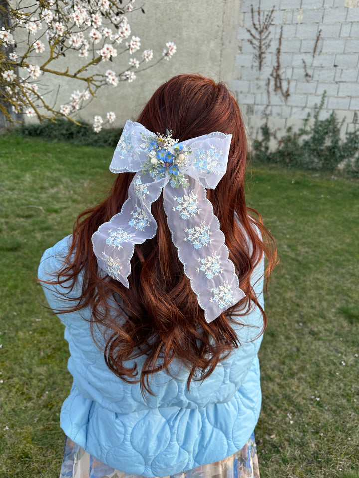 A person outdoors on grass wearing a light blue quilted jacket and a long, wavy reddish-brown hair accessory with a sheer white lace ribbon and white floral applique.