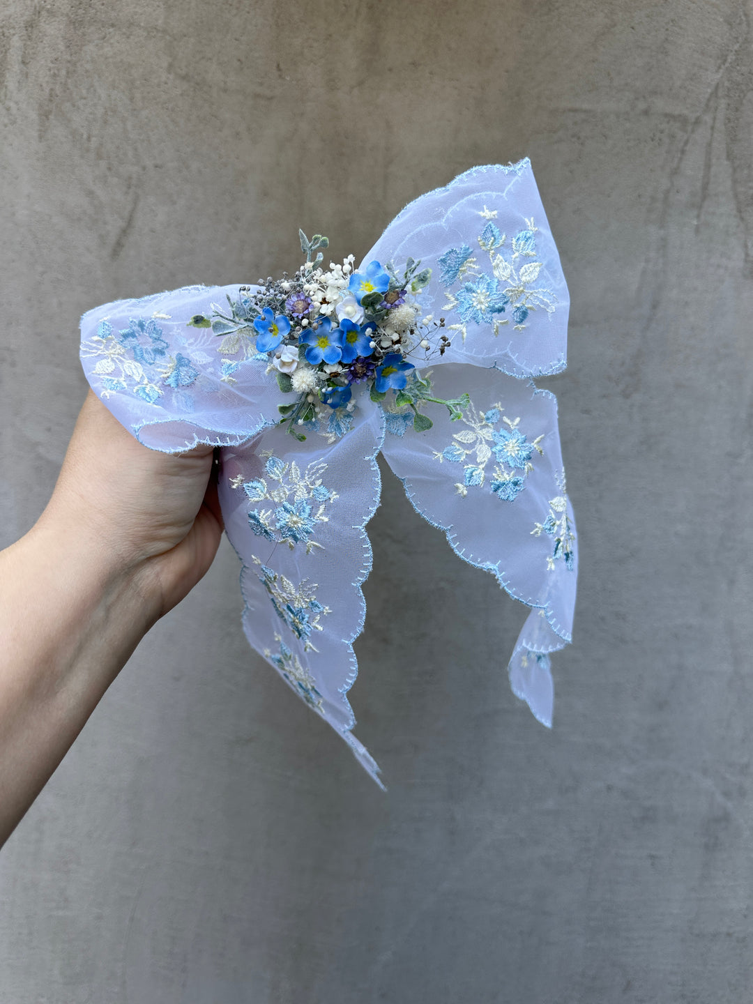 A hand holding a light blue embroidered fabric flower hair bow with blue and white floral details against a textured gray background.