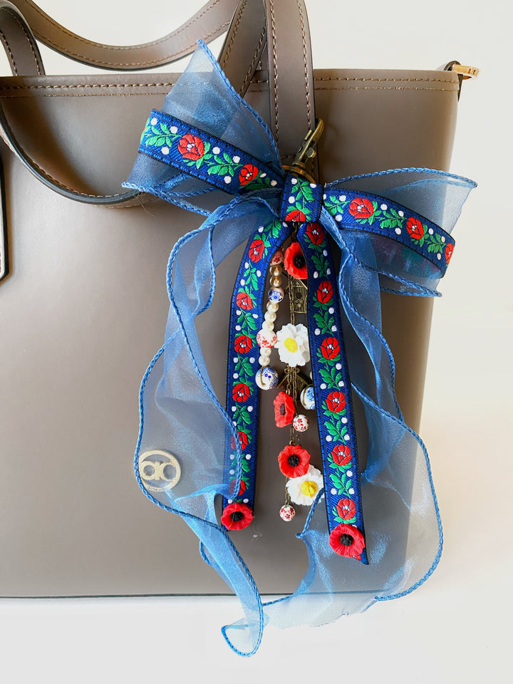 A light brown leather tote bag decorated with a large blue sheer ribbon bow featuring colorful floral embroidery and hanging charms.