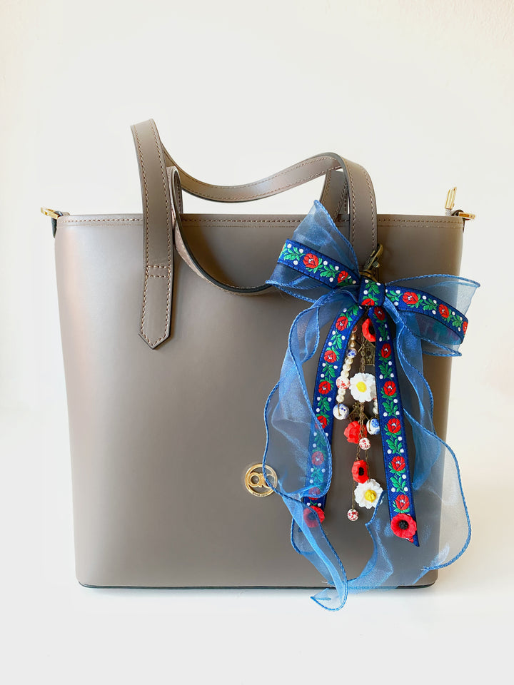 A taupe leather tote bag with a large blue sheer organza bow decorated with colorful embroidered flowers and beads, shown against a plain white background.