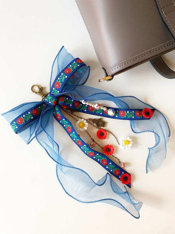 A blue sheer ribbon keychain or bag charm with a floral pattern, gold clasp, and decorative flower charms, styled next to a taupe leather bag on a white background.
