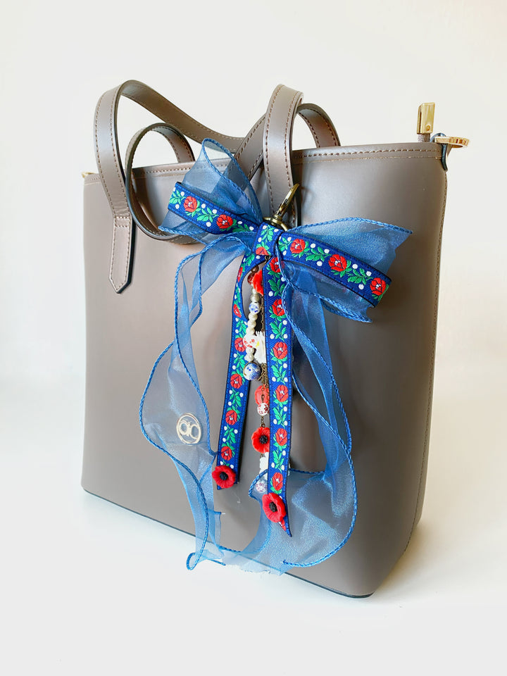 A taupe faux-leather handbag with dual top handles, a gold zipper, and a large blue sheer ribbon bow decorated with a colorful floral pattern and red pom-poms, shown on a white background.