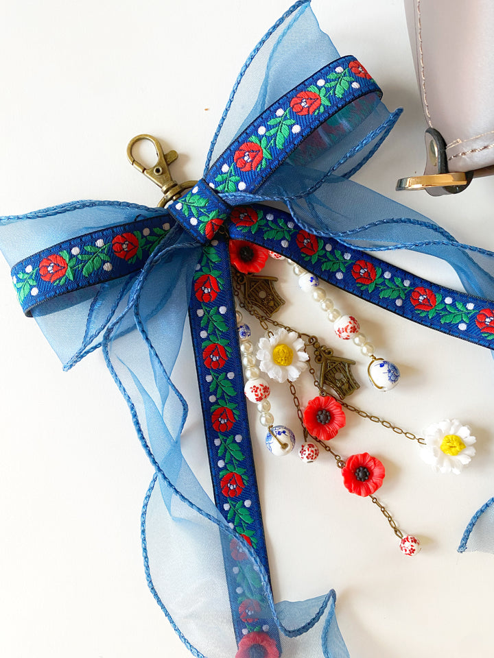 A blue sheer ribbon bow with a floral pattern, attached to a gold clasp and decorated with small red poppy flowers, white daisy beads, and brown woven accents on a white background.
