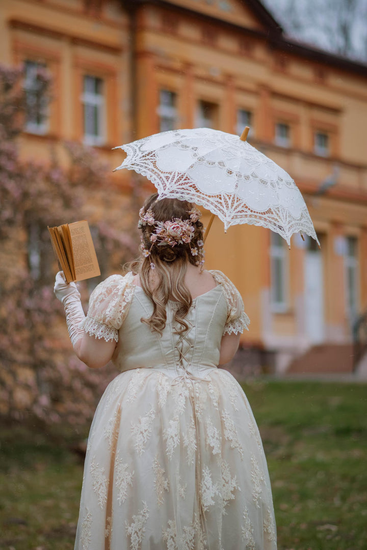 Woman in a vintage lace dress holding a lace umbrella and book, standing in front of a yellow building.