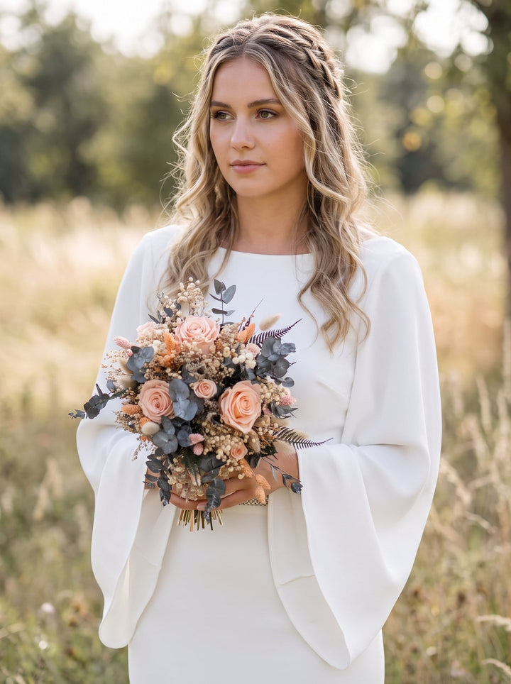 A woman standing outdoors in a field wearing a white bridal dress with dramatic flared sleeves and holding a bouquet of peach, blush, and muted blue-gray flowers.