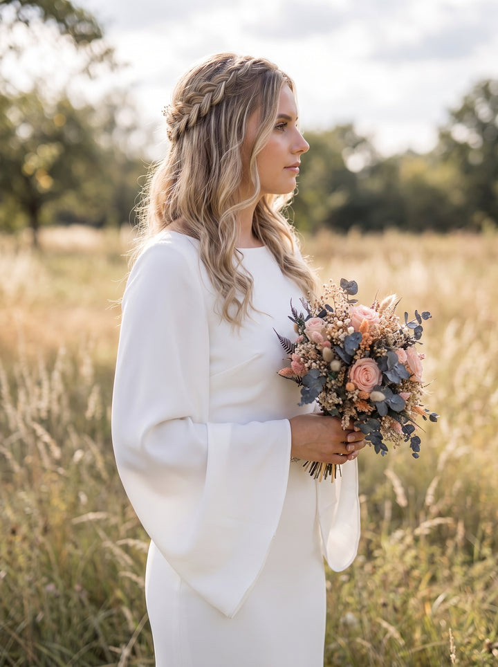 A woman in a white long-sleeve wedding dress stands in a sunlit grassy field, holding a bouquet of blush pink, peach, and muted blue flowers.