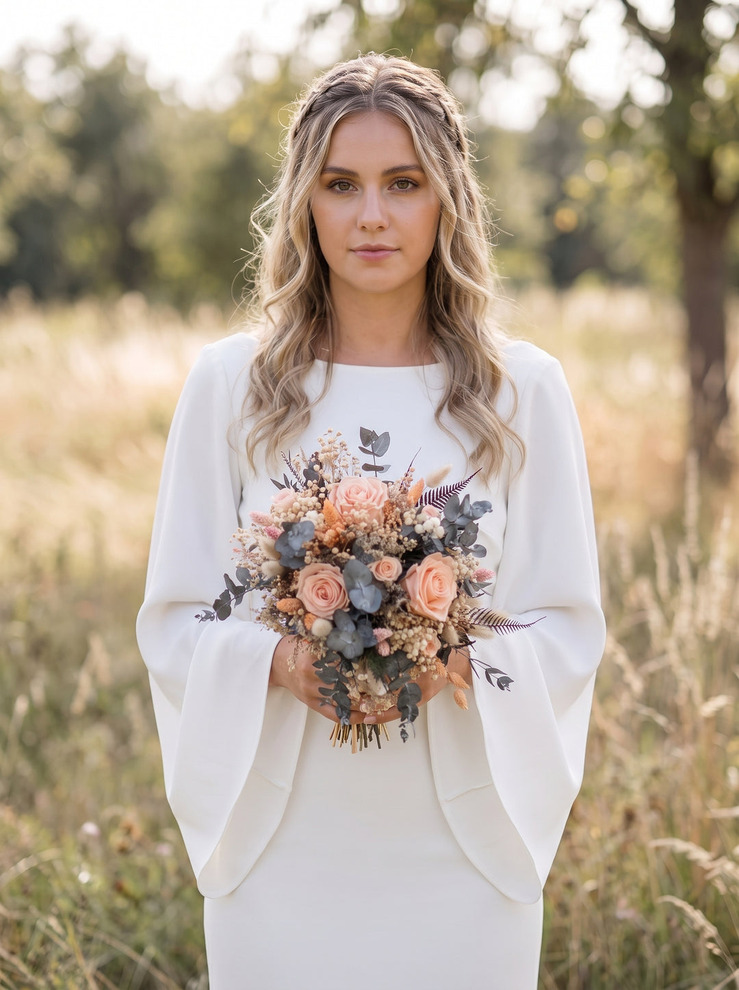 A woman standing outdoors in a field wearing a white long-sleeve dress with a delicate butterfly print on the chest and holding a peach and gray floral bouquet.