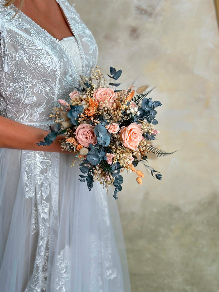 A close-up of a bride in a white lace wedding dress holding a dried floral bouquet with blush pink roses, dusty blue eucalyptus, and beige filler flowers against a soft beige textured background.