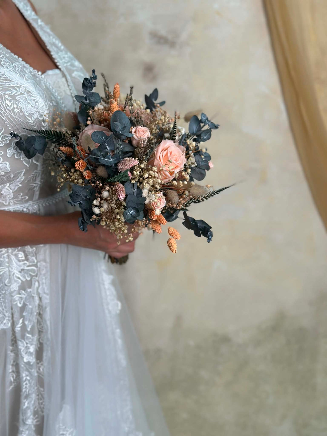 A close-up of a person in a white lace wedding dress holding a dried floral bouquet with dusty pink roses, muted blue-green foliage, and peach dried accents against a soft beige wall.
