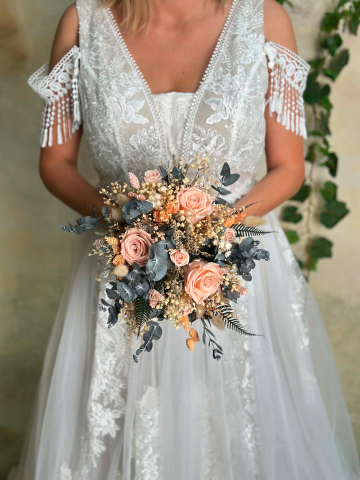 A bride wearing a white lace and tulle wedding dress with cold-shoulder fringe sleeves, holding a dried floral bouquet with peach roses, blue-gray foliage, and beige filler flowers.