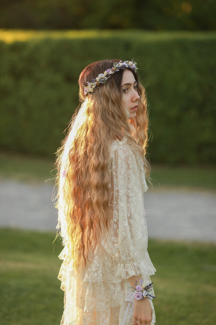 Woman with long hair and floral crown standing outdoors with greenery in the background