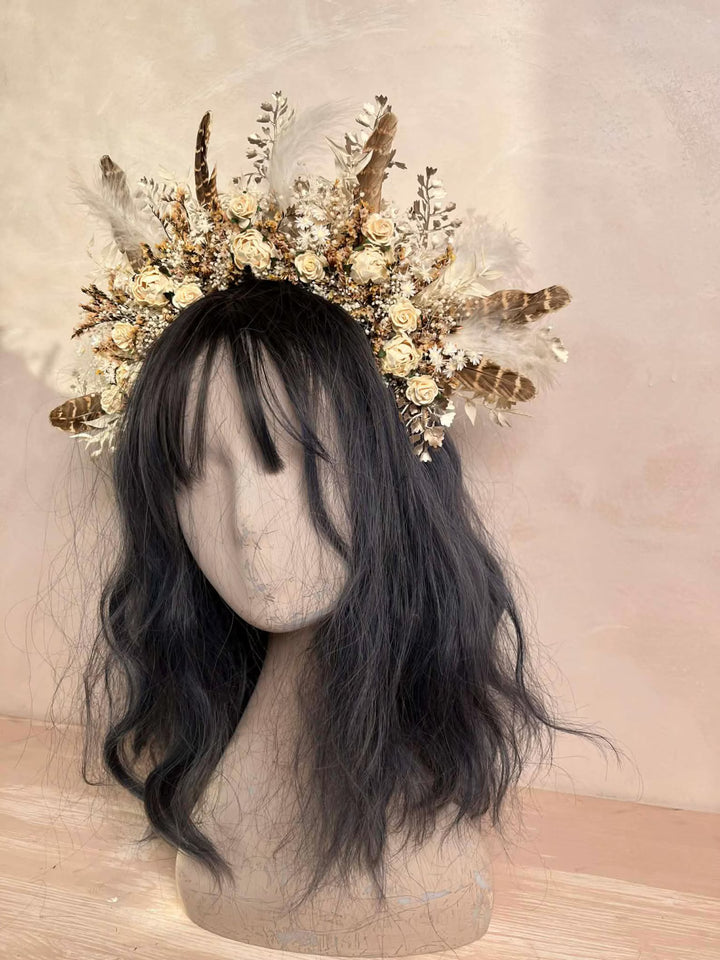 Mannequin head with black hair wearing a decorative headpiece with feathers and flowers in earthy tones on a beige background.
