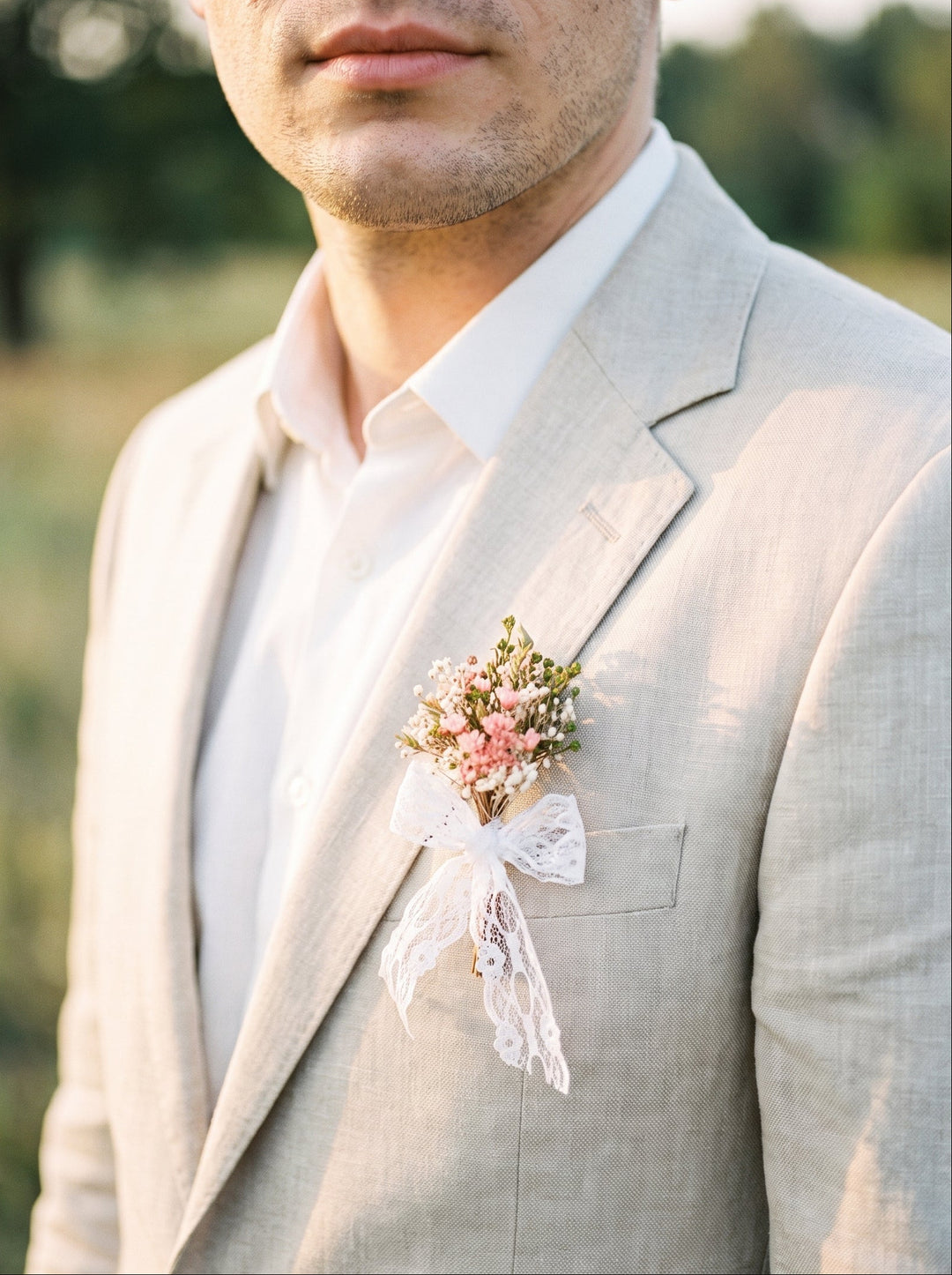 A man wearing a light beige linen suit jacket over a white dress shirt, featuring a delicate floral boutonniere with pink and white flowers and a white lace ribbon on his lapel, set against a softly blurred outdoor background.