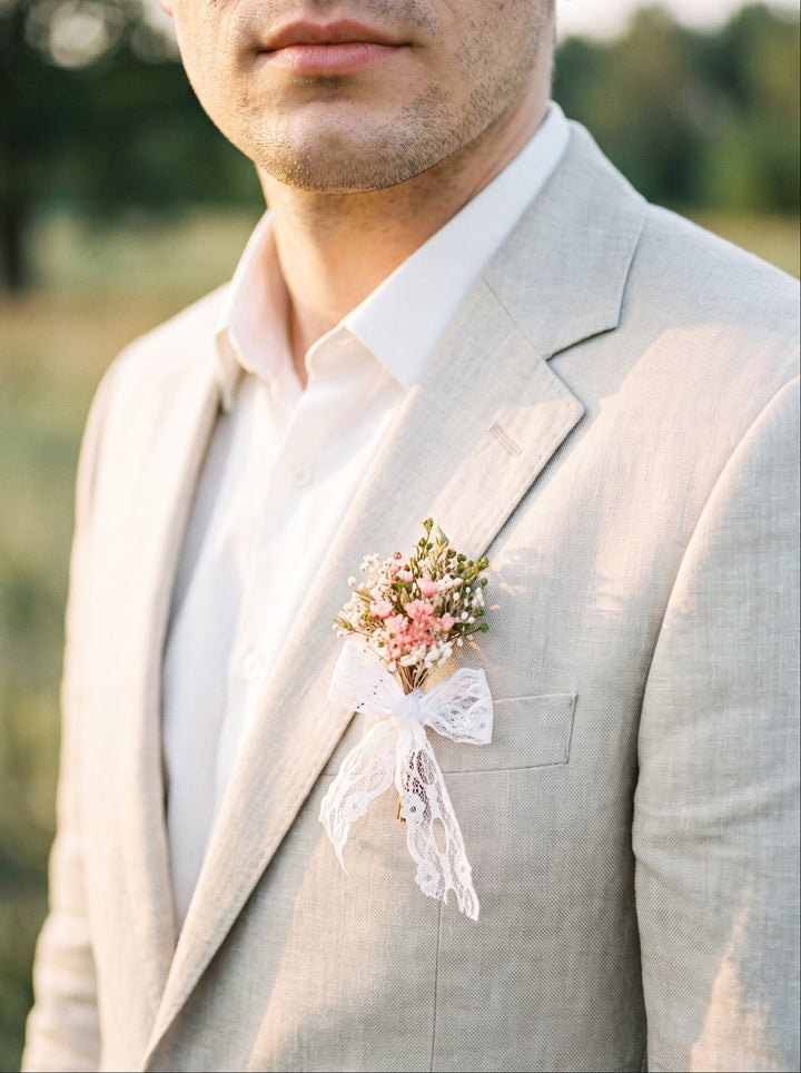 A man wearing a light beige linen suit jacket over a white dress shirt, featuring a delicate floral boutonniere with pink and white flowers and a white lace ribbon on his lapel, set against a softly blurred outdoor background.
