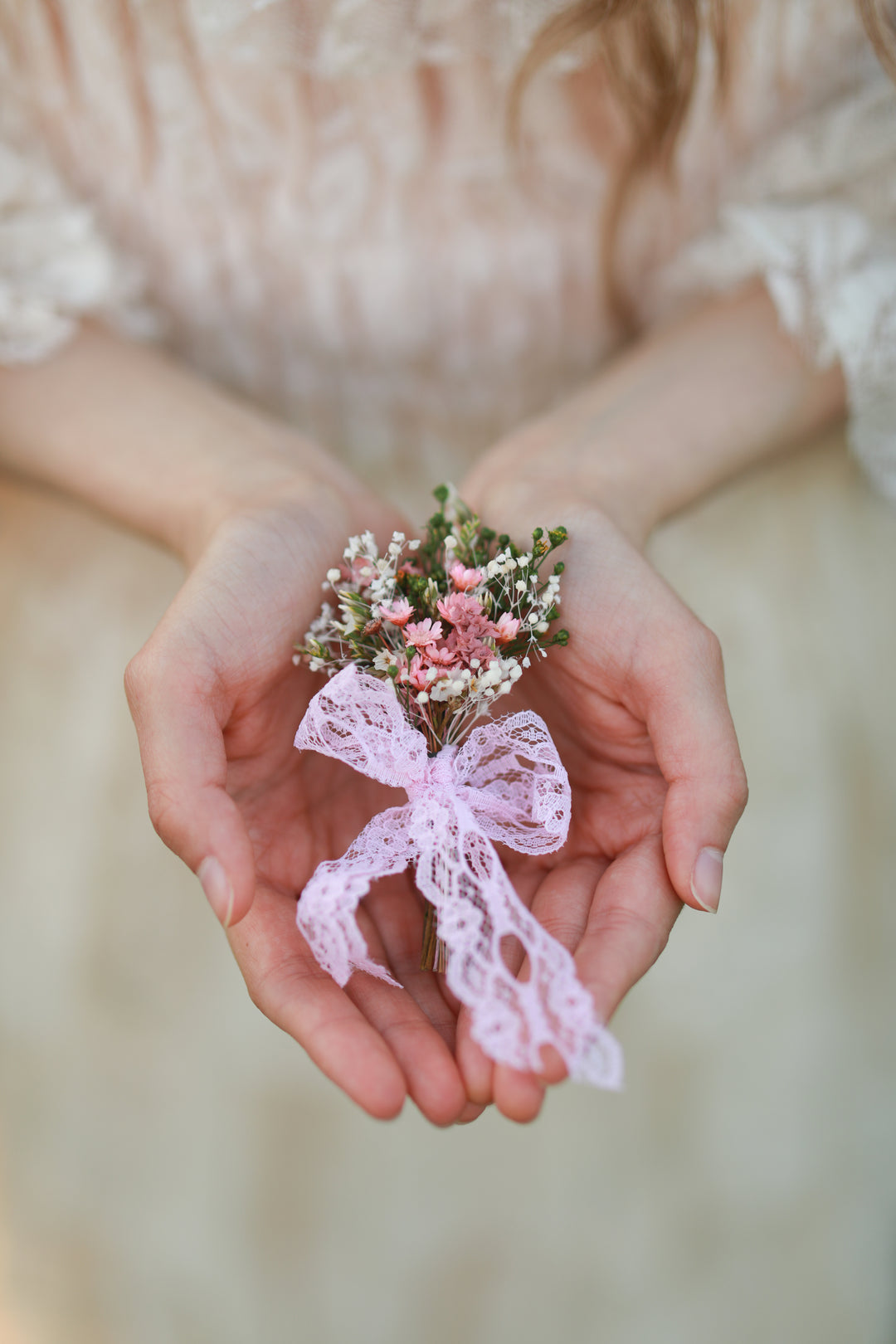 A close-up of a person's hands holding a small floral boutonniere with pink and white flowers tied with a light pink lace ribbon, against a soft beige dress background.