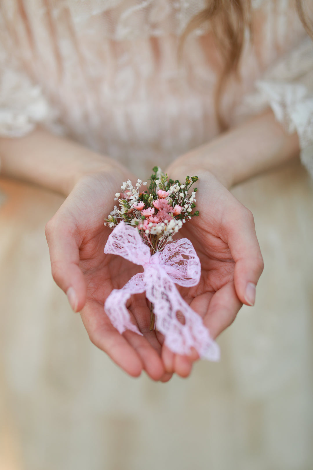 A delicate floral boutonniere with pink blossoms, white baby's breath, and a light pink lace ribbon bow, held gently in a person's hands against a soft beige dress background.