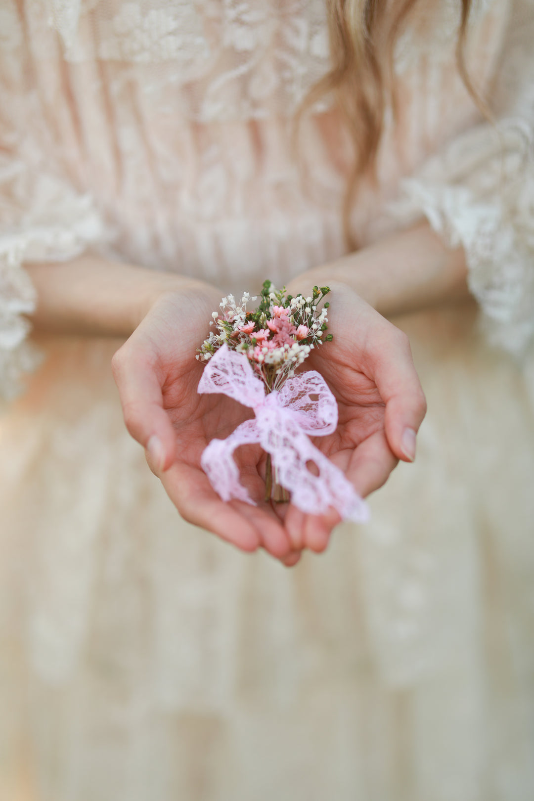 A delicate floral boutonniere with small pink and white flowers tied with a light pink lace ribbon, held in a person's hands wearing a soft cream lace dress.
