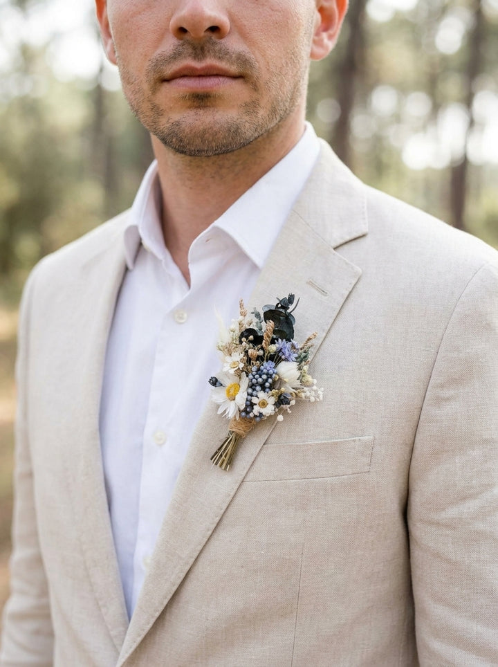 A man wearing a light beige linen suit jacket over a white dress shirt, featuring a delicate dried flower boutonniere on the chest pocket, set against a soft, blurred forest background.