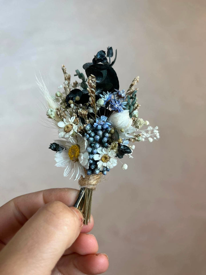 Handcrafted boutonniere made entirely from preserved and dried botanicals in a soft, warm palette - a central ivory daisy, delicate ixodia clusters, silvery eucalyptus sprigs, and wispy dried grasses.