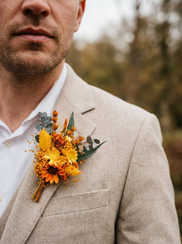 A close-up of a man wearing a light beige linen suit jacket over a white collared shirt, featuring a vibrant orange and yellow floral boutonniere on his lapel against a soft, blurred outdoor background.