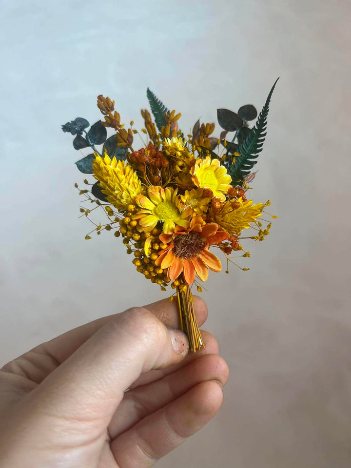 A hand holding a small flower boutonniere with yellow and orange flowers, green leaves, and gold wrapping on the stems, set against a plain light gray background.