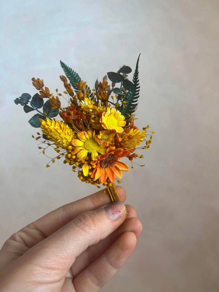 A hand holding a small floral boitonniere with yellow and orange flowers, brown filler stems, and green fern-like leaves against a soft beige background.