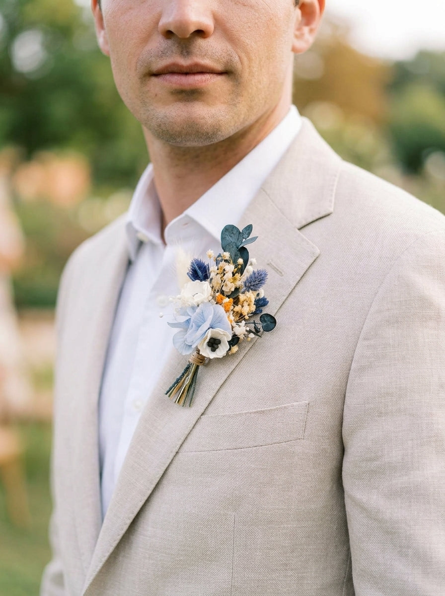 A close-up of a man wearing a light beige linen suit jacket, white dress shirt, and a delicate dried flower boutonniere with blue, white, and gold tones on his lapel, set against a soft, blurred outdoor background.