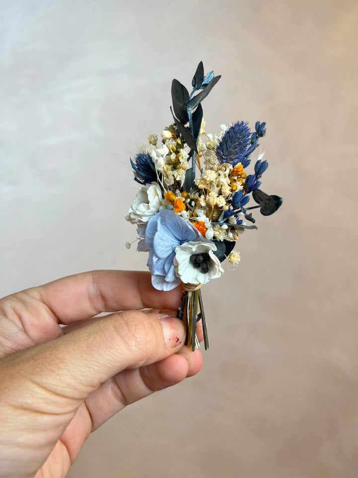 A hand holding a boutonniere with blue, cream, white, and orange botanical elements against a soft beige background.