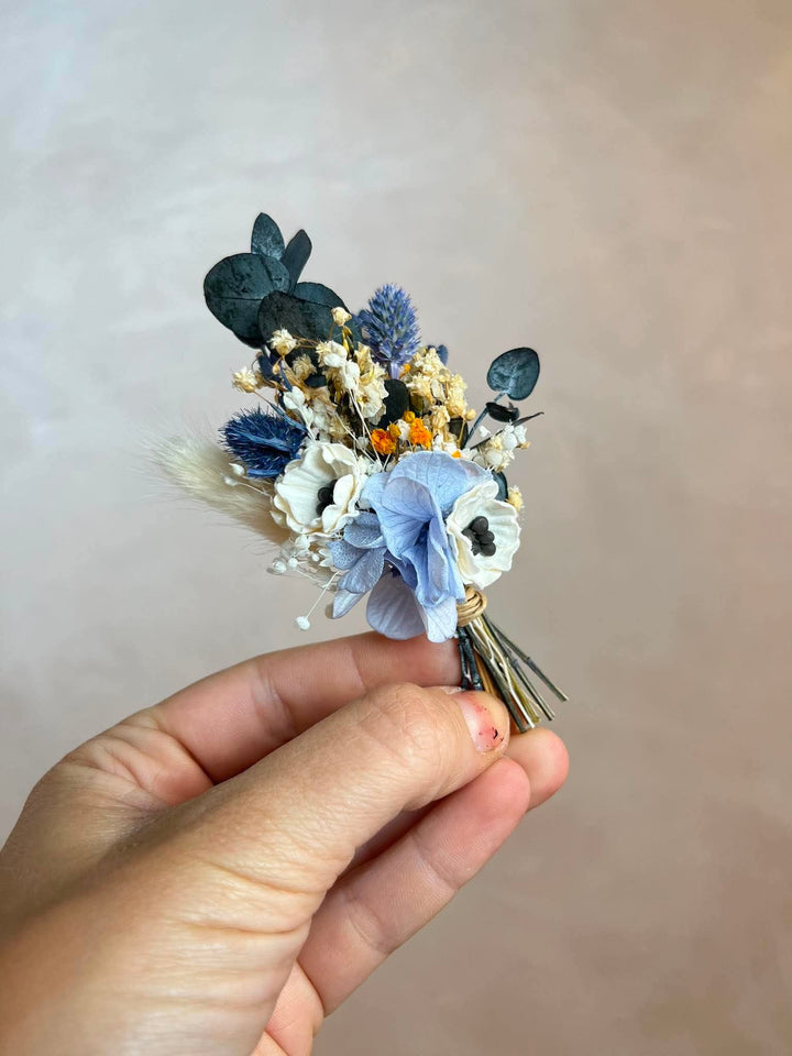 A hand holding a small flower boutonniere with light blue, white, navy, and beige botanical elements against a soft beige background.
