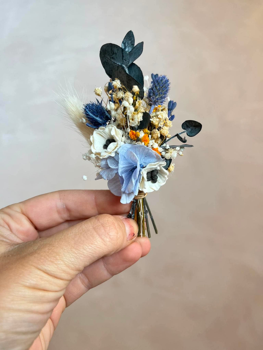 A hand holding a small flower corsage with anemone, blue lavender, dark eucalyptus leaves, and baby's breath against a soft beige background.