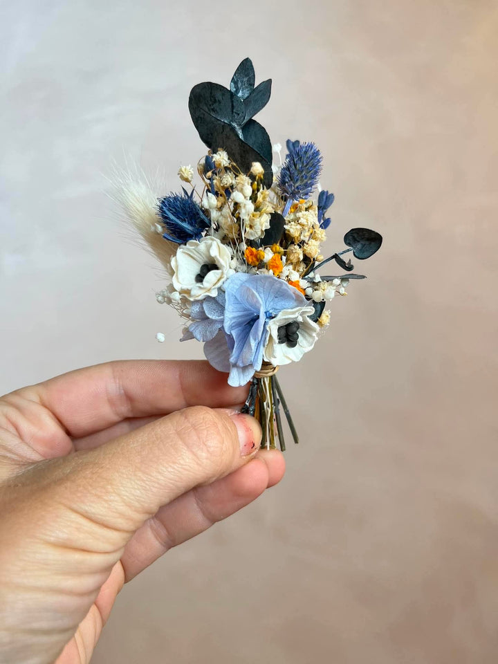 A hand holding a small flower corsage with anemone, blue lavender, dark eucalyptus leaves, and baby's breath against a soft beige background.