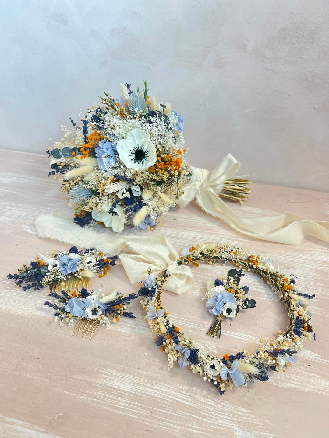 A coordinated floral set arranged on a pale wood surface against a soft neutral wall, including a bouquet, a headpiece, a wrist/forehead piece, and a circular wreath. The florals are primarily cream, dusty blue, navy, white, and small orange accents.