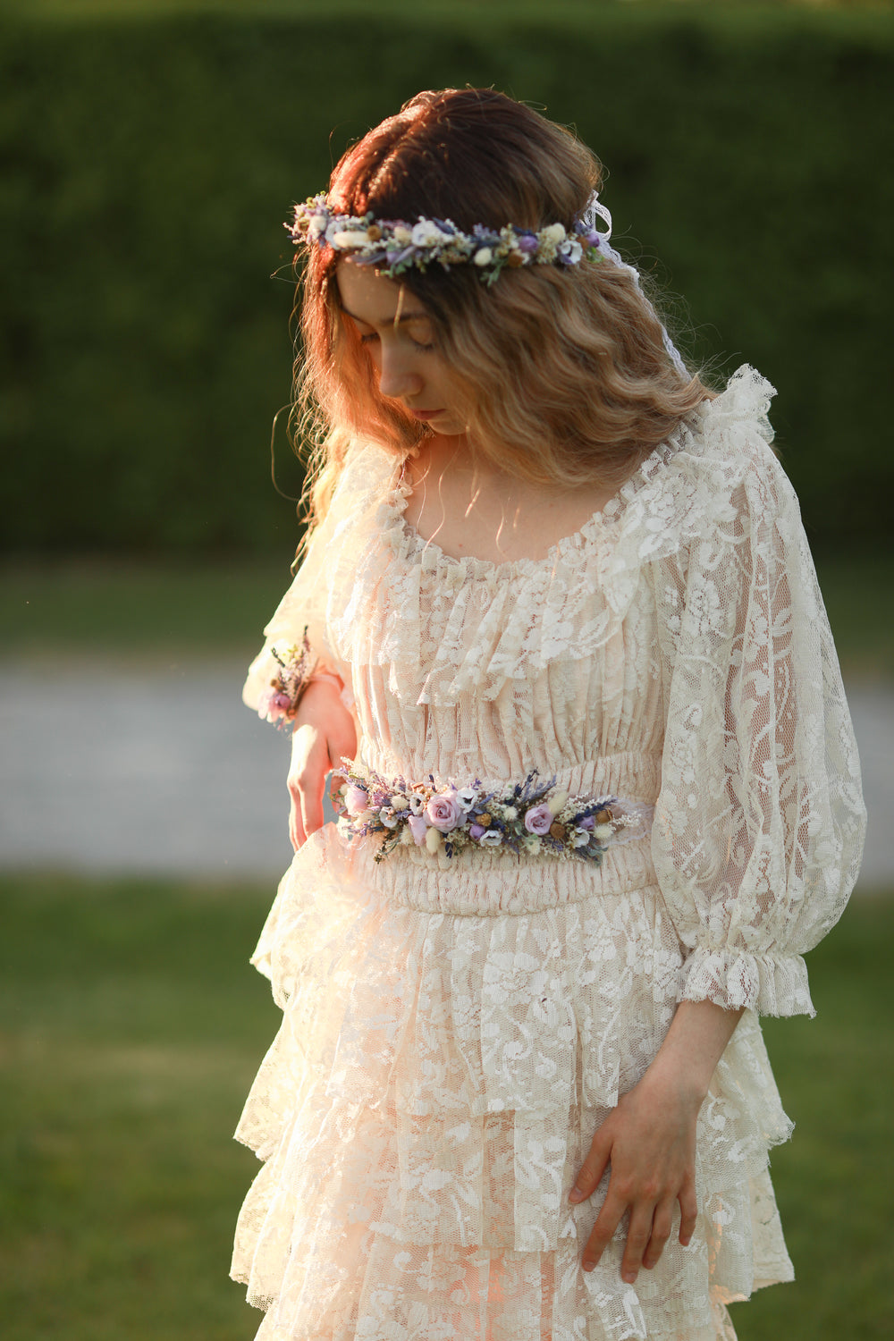 Woman in a white lace dress with floral accessories standing outdoors.