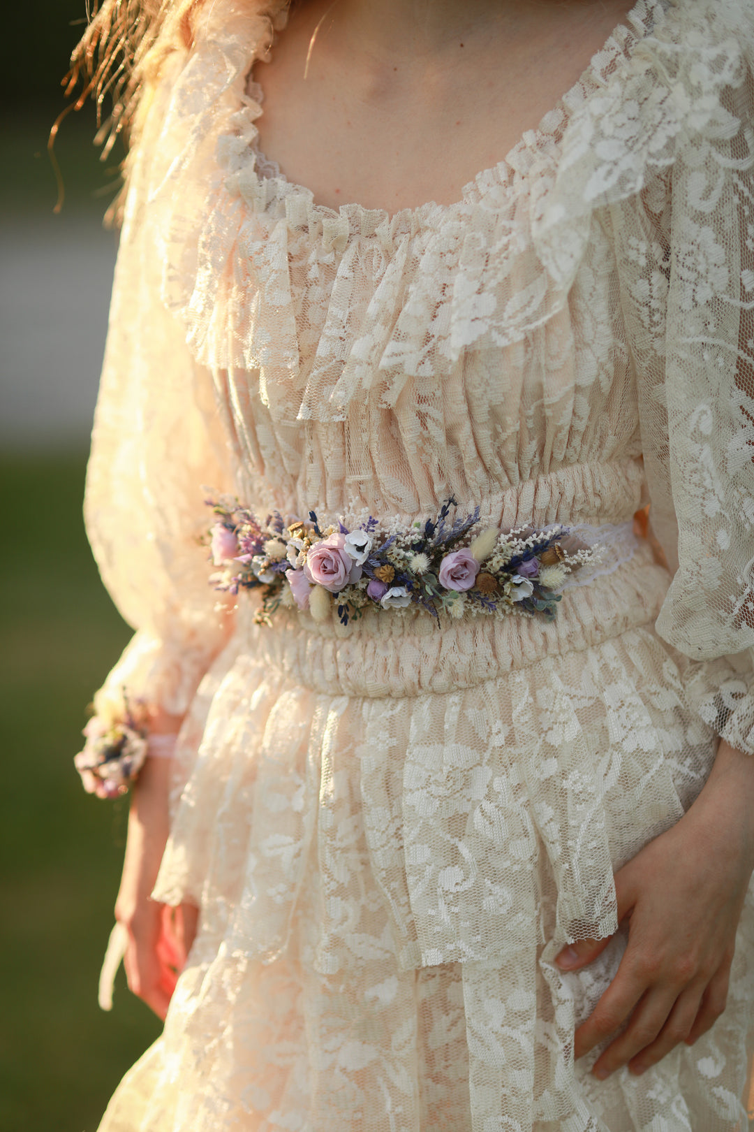 Close-up of a person wearing a lace dress with a floral belt against a blurred natural background