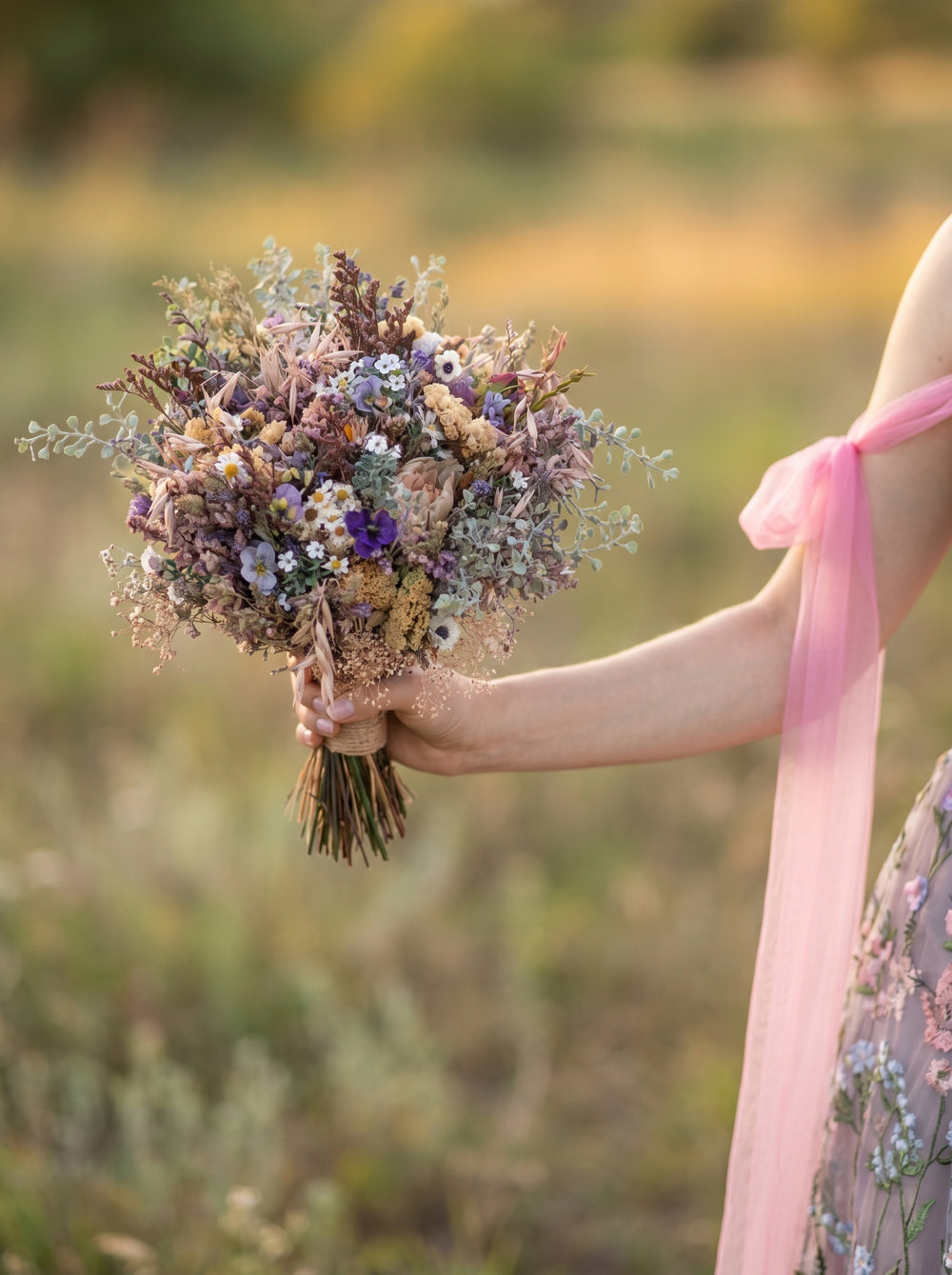Person holding a bouquet of purple, blue and cream flowers in a field