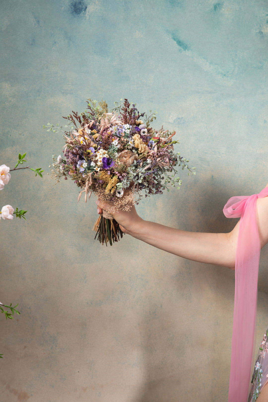 Hand holding a bouquet of purple, blue, cream and blush flowers against a textured gray background