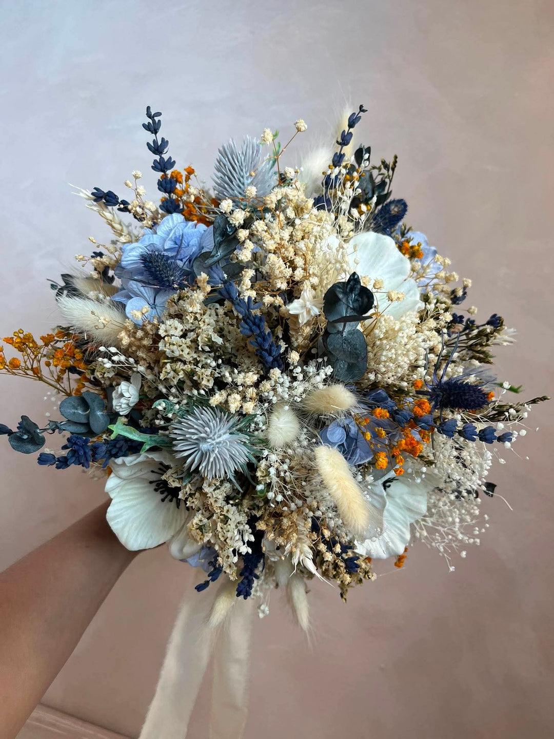 A flower bouquet held by a hand, featuring light blue hydrangeas, white baby's breath, navy blue thistles, beige pampas grass, and small orange accents against a soft beige background.