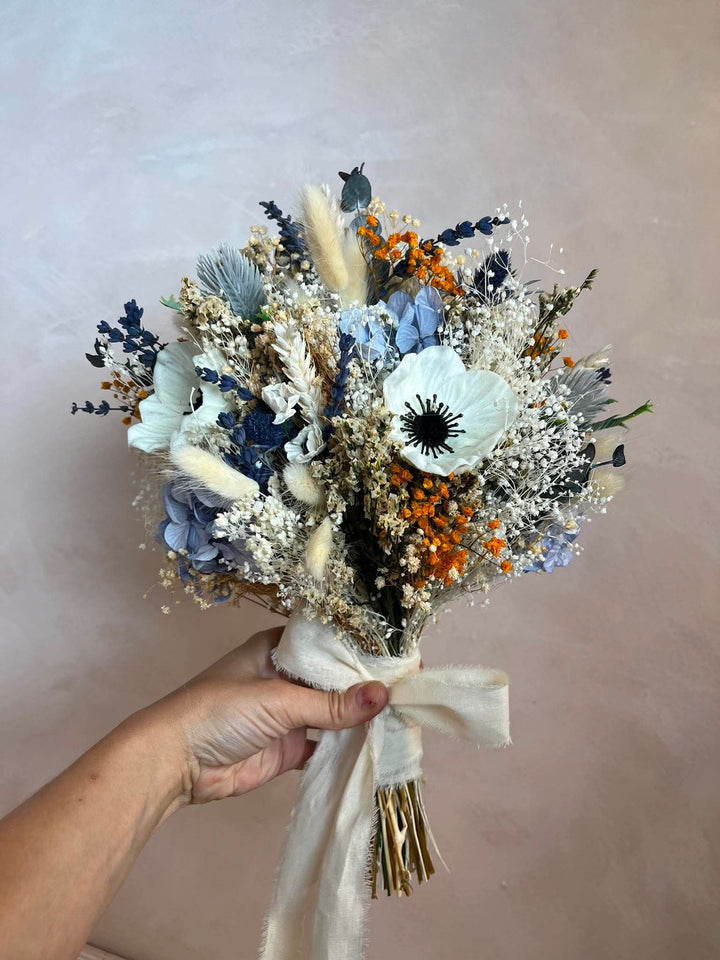 A flower bouquet held by a hand, featuring light blue hydrangeas, white anemones, blue pampas grass, orange berries, and a cream ribbon bow, set against a soft beige background.