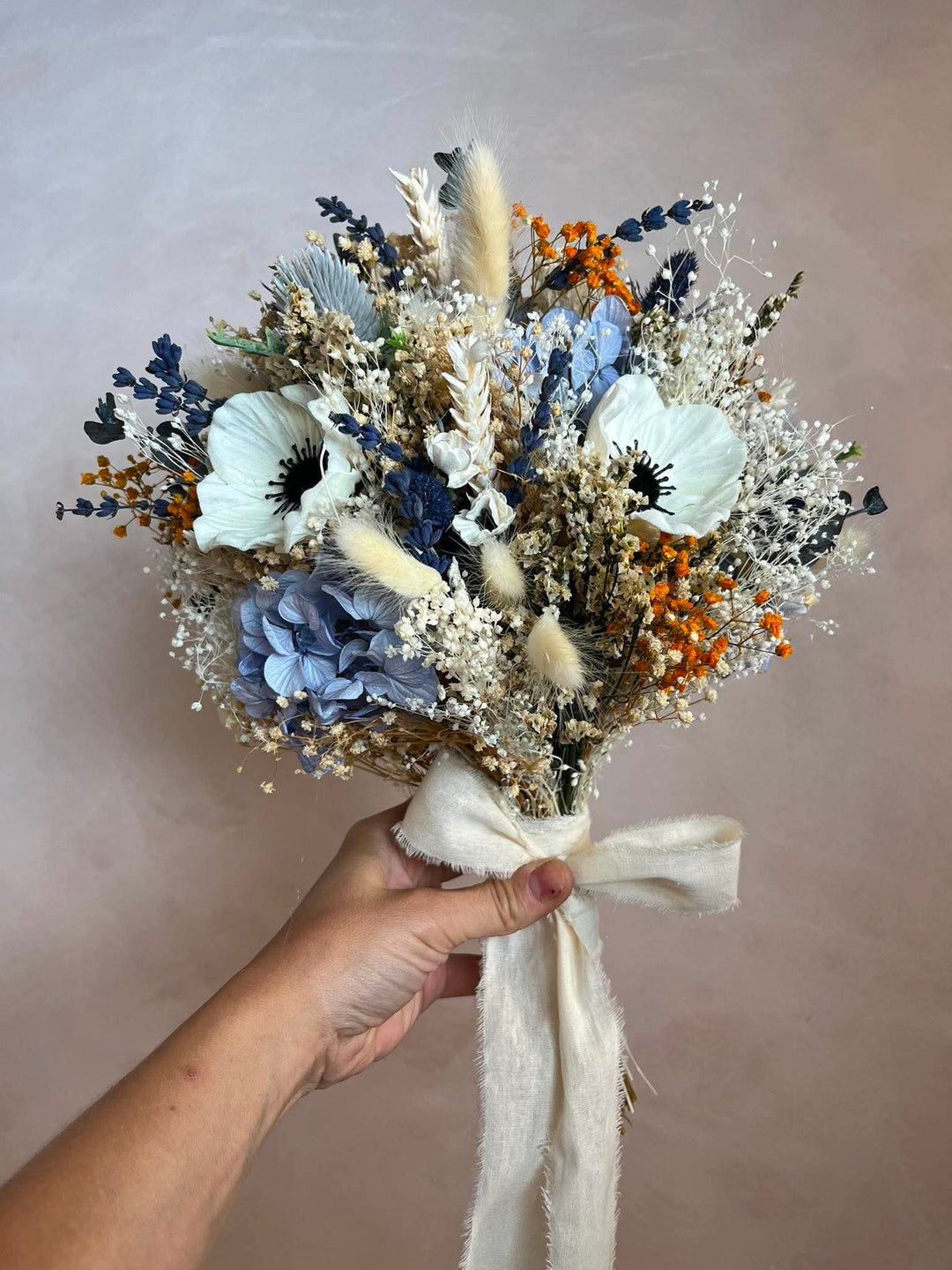 A hand holding a flower bouquet featuring white anemones, light blue hydrangeas, orange berries, and beige pampas grass, tied with a cream ribbon against a soft beige background.