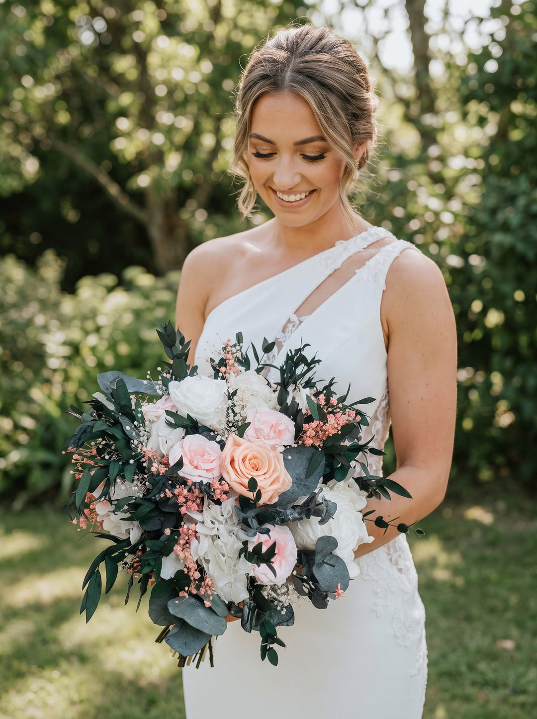 A smiling bride in a white one-shoulder wedding dress holds a large bridal bouquet with white and blush pink roses, green foliage, and small pink berries, standing outdoors in front of a blurred green garden background.