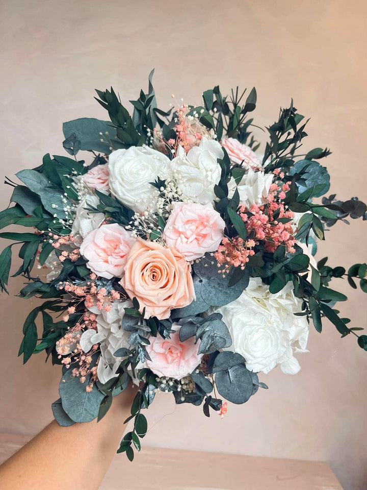A bouquet of preserved flowers held by a hand, featuring white and peach roses, dusty pink filler blooms, and green eucalyptus foliage against a soft beige background.