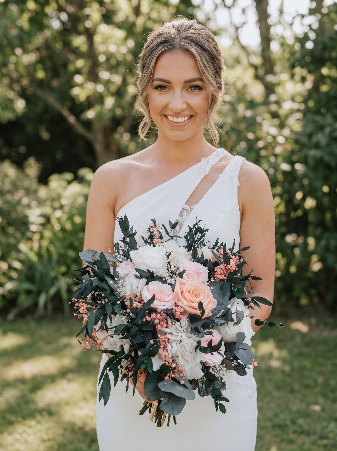 A smiling bride in a white one-shoulder wedding dress holds a large bridal bouquet with pink roses, white flowers, and green foliage, standing outdoors in front of a blurred green garden background.