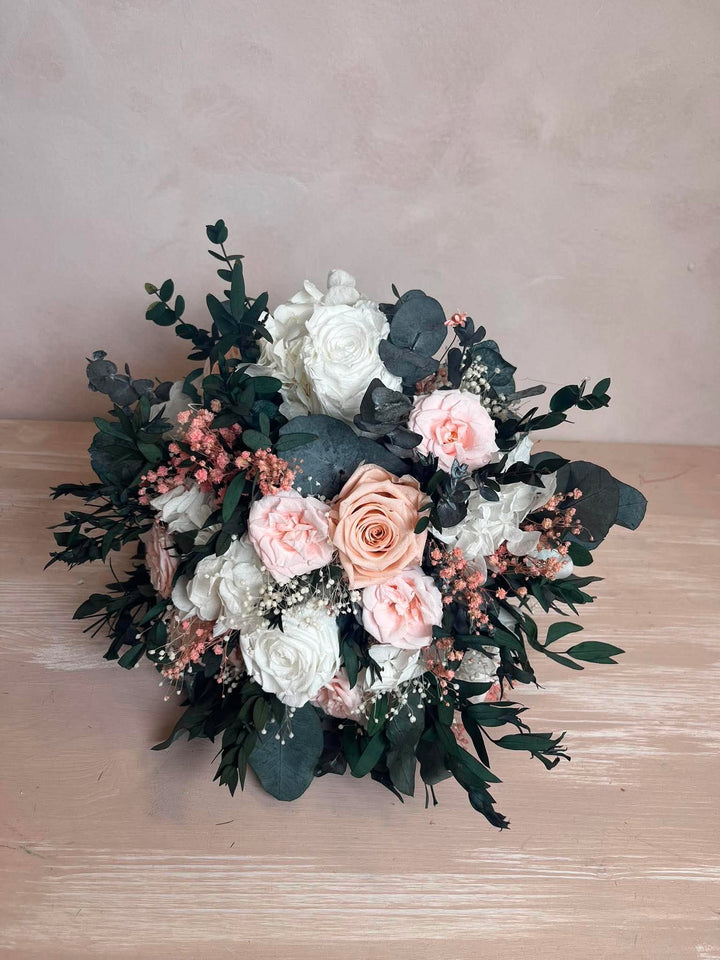 A round floral arrangement featuring white and blush pink roses, green eucalyptus leaves, and small flowers, displayed on a light wood surface against a textured beige wall.